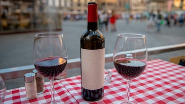 eine Flasche Rotwein und zwei gefüllte Weingläser mit Rotwein auf einem Tisch an einer Piazza, die unscharf im Hintergrund zu sehen ist; | Bild: 	mauritius images / TravelFlow / Alamy / Alamy Stock Photos