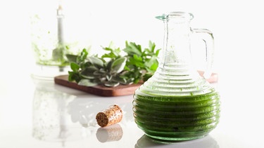 Eine Glaskaraffe mit grünem Kräuteröl; daneben liegen Kräuter. | Bild: 	mauritius images / foodcollection